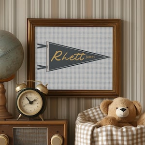 Puede incluir: Un banderín enmarcado con el nombre "Rhett James" cuelga sobre una radio vintage, un despertador y un globo terráqueo. Un osito de peluche descansa en una cesta forrada con cuadros. Los objetos están colocados sobre una cómoda blanca.