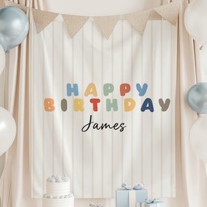 Peut inclure: Une toile de fond d'anniversaire avec des rayures verticales beiges, une inscription "HAPPY BIRTHDAY" aux lettres arrondies colorées et le prénom "James" en écriture noire. L'ensemble comprend un gâteau blanc, des cadeaux et des ballons.