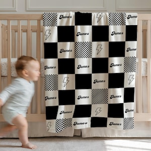 Può includere: Una coperta per bambini a scacchi bianchi e neri con il nome "James" stampato sopra. La coperta presenta anche grafiche a forma di fulmine. La coperta è drappeggiata su una culla di legno.