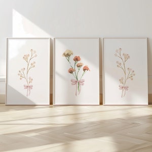 Könnte beinhalten: Drei gerahmte Aquarelldrucke von Blumensträußen mit rosa Schleifen. Die Sträuße zeigen zarte weiße Blüten und orangefarbene Blüten.