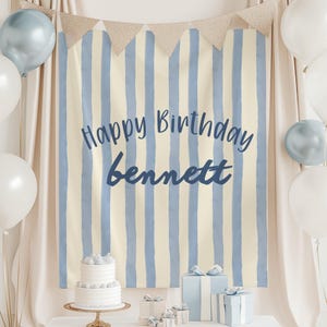 Può includere: Uno sfondo rettangolare in tessuto con strisce verticali color crema e azzurro, recante la scritta Happy Birthday bennett in corsivo blu scuro. Lo sfondo è decorato con una ghirlanda di bandierine e circondato da palloncini bianchi e azzurri.