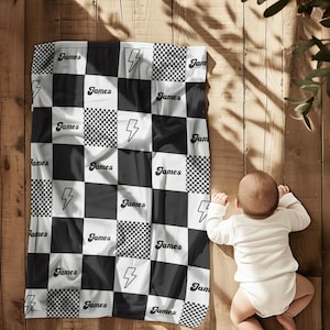 Puede incluir: Una manta de bebé a cuadros blancos y negros con el nombre "James" y gráficos de rayos. La manta está sobre una superficie de madera. Un bebé con un body blanco está acostado al lado.