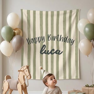 May include: A fabric backdrop featuring vertical sage green and cream stripes with the text Happy Birthday luca in dark gray script. The scene includes clusters of sage green, cream, and brown balloons, a wooden rocking horse, and gift boxes.