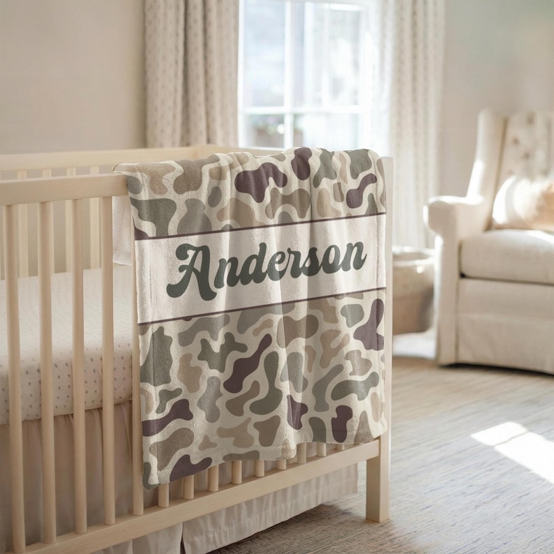 May include: A soft blanket featuring a repeating organic camouflage pattern in shades of beige, tan, and dark brown. A horizontal white band runs across the center, displaying the name Anderson in a dark gray, cursive, retro-style font.