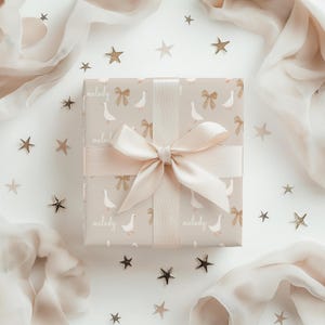 May include: A gift box wrapped in beige paper with a white goose and bow pattern. The box is tied with a light pink satin ribbon and bow. The paper has the word "melody" printed on it. The box is surrounded by gold stars on a white background.
