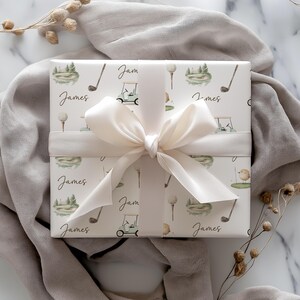 May include: A gift wrapped in paper with a golf-themed pattern. The paper is decorated with golf clubs, golf carts, and golf balls. The name "James" is written on the paper. A cream ribbon and bow are tied around the gift, set against a neutral backdrop.