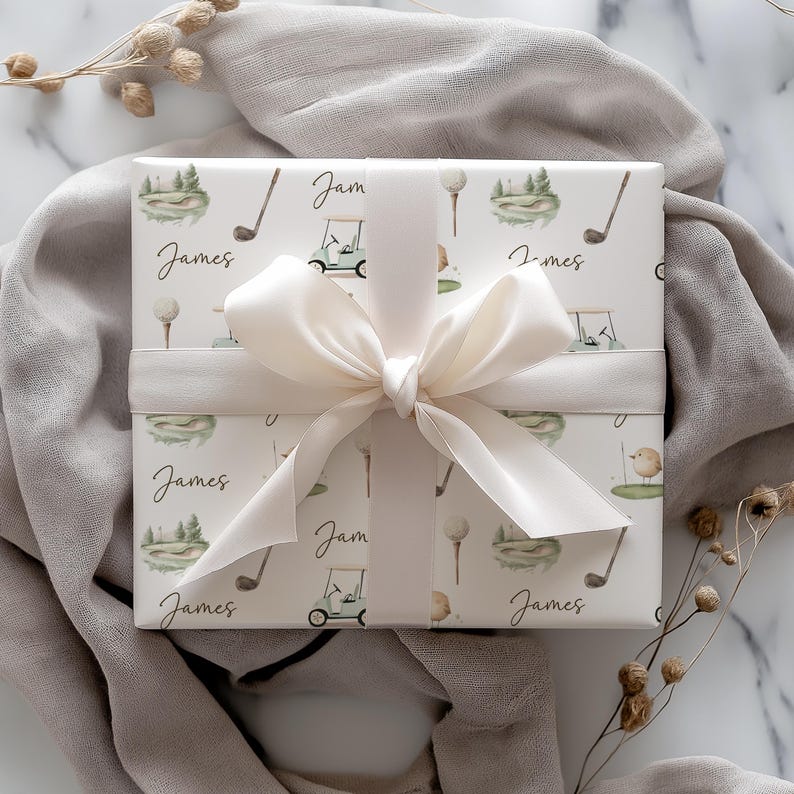 May include: A gift wrapped in paper with a golf-themed pattern. The paper is decorated with golf clubs, golf carts, and golf balls. The name "James" is written on the paper. A cream ribbon and bow are tied around the gift, set against a neutral backdrop.