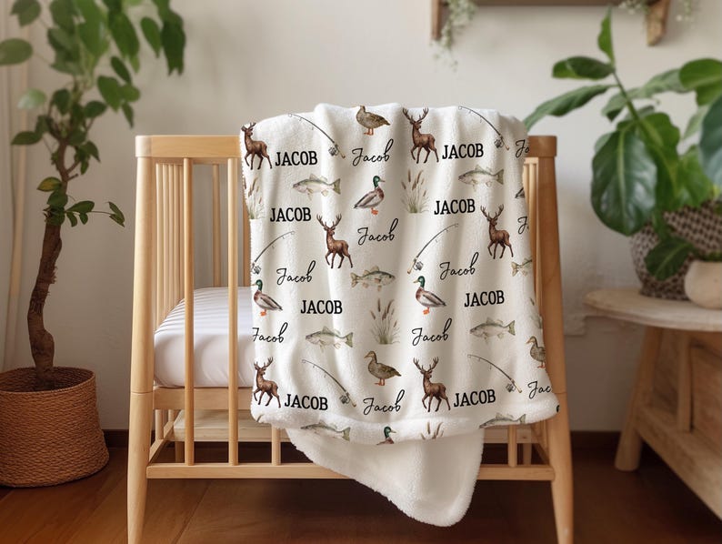 May include: A cream-colored baby blanket with a repeating pattern of deer, fish, ducks, and fishing rods. The name "Jacob" is written in a script font throughout the design. The blanket is draped over a wooden crib.