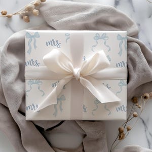 K&ouml;nnte beinhalten: Wei&szlig;e Geschenkpapier mit einem blauen und wei&szlig;en Schleifenmuster und dem Wort "mrs" wiederholt sich &uuml;berall. Das Geschenk ist mit einem wei&szlig;en Satinband verpackt.