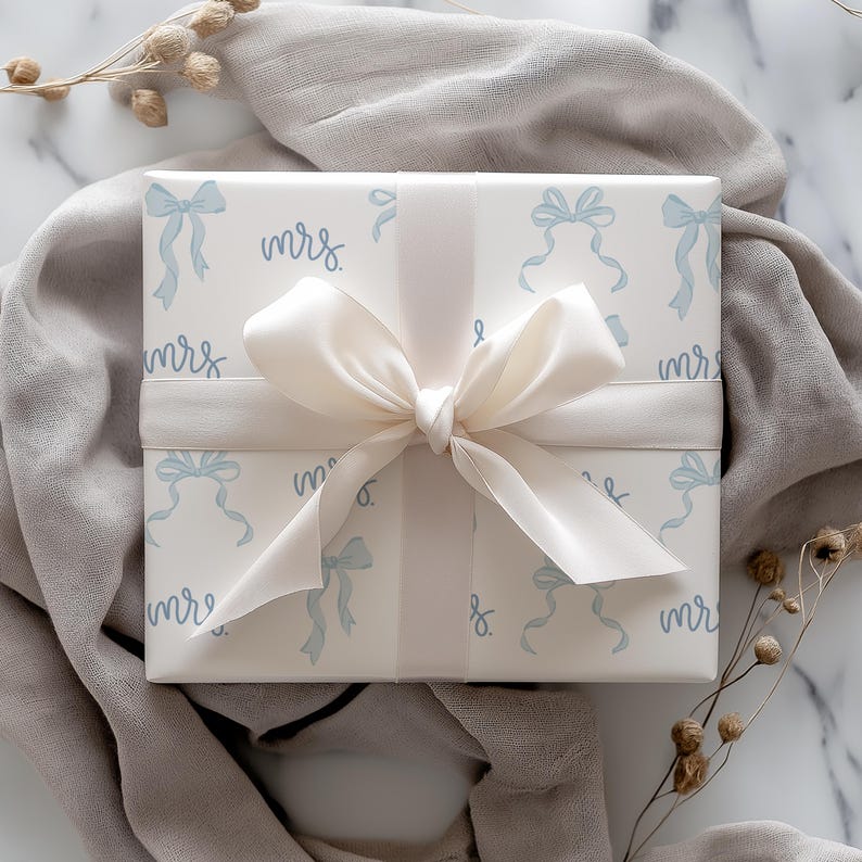 K&ouml;nnte beinhalten: Wei&szlig;e Geschenkpapier mit einem blauen und wei&szlig;en Schleifenmuster und dem Wort "mrs" wiederholt sich &uuml;berall. Das Geschenk ist mit einem wei&szlig;en Satinband verpackt.