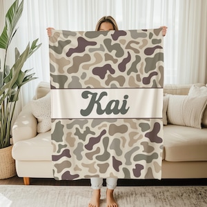 May include: A rectangular throw blanket featuring a camouflage pattern in shades of beige, tan, and dark brown. A horizontal white band across the center displays the name Kai in a dark brown, stylized, cursive-inspired font.