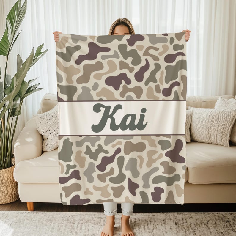 May include: A rectangular throw blanket featuring a camouflage pattern in shades of beige, tan, and dark brown. A horizontal white band across the center displays the name Kai in a dark brown, stylized, cursive-inspired font.