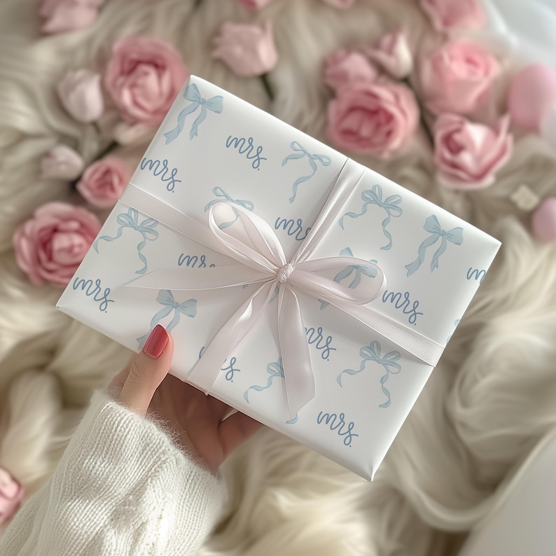 K&ouml;nnte beinhalten: Wei&szlig;e Geschenkpapier mit einem blauen und wei&szlig;en Schleifenmuster. Das Papier ist mit dem Wort "mrs." durchgehend bedruckt.