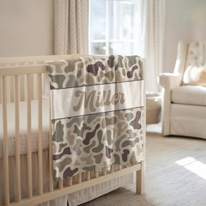 May include: A soft baby blanket featuring a camouflage pattern in shades of beige, tan, and dark brown. A horizontal white band runs across the center of the blanket, displaying the name Miller in a brown, cursive, script-style font.