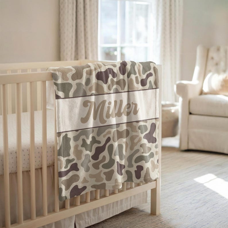 May include: A soft baby blanket featuring a camouflage pattern in shades of beige, tan, and dark brown. A horizontal white band runs across the center of the blanket, displaying the name Miller in a brown, cursive, script-style font.