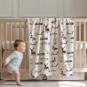 May include: A cream-colored baby blanket with a repeating pattern of deer, fish, fishing poles, and ducks. The name "Jacob" is printed in a bold font, and the name "Jacob" is also written in a script font. The blanket is draped over a wooden crib.