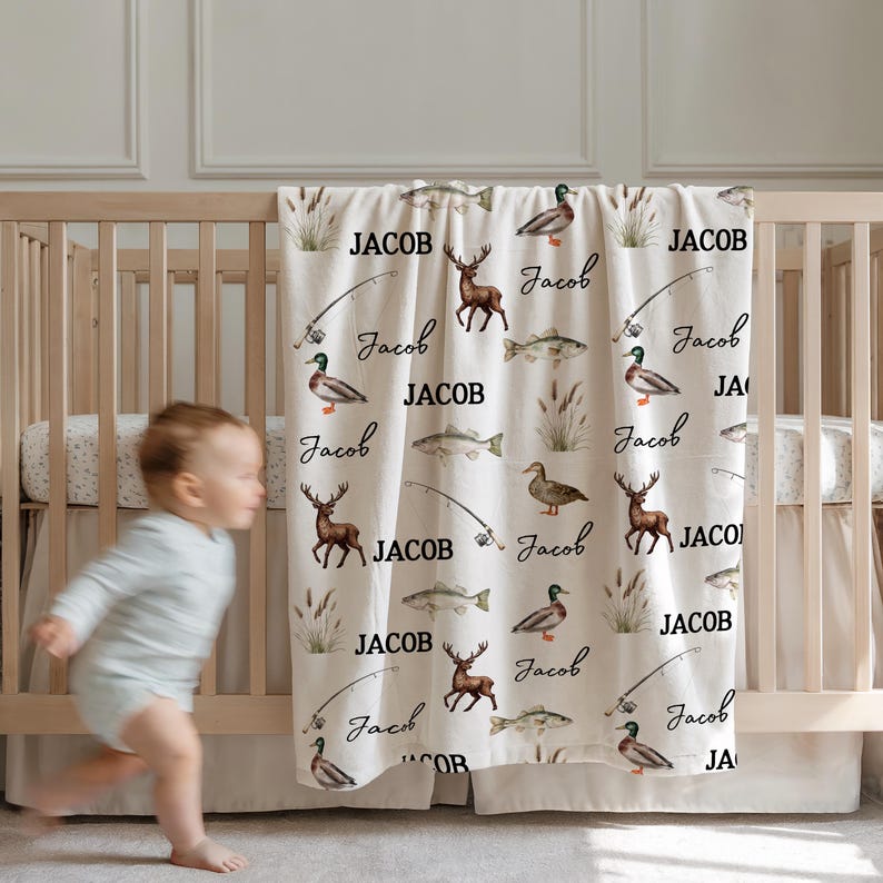 May include: A cream-colored baby blanket with a repeating pattern of deer, fish, fishing poles, and ducks. The name "Jacob" is printed in a bold font, and the name "Jacob" is also written in a script font. The blanket is draped over a wooden crib.