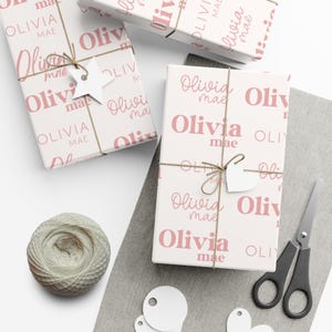May include: Gift boxes wrapped in white paper with a repeating pattern of the name "Olivia Mae" in pink. Each box is tied with twine and a white tag. A ball of twine, scissors, and blank tags are also present.