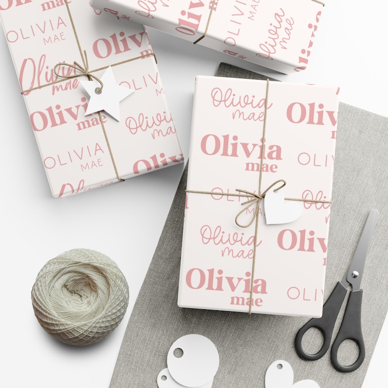May include: Gift boxes wrapped in white paper with a repeating pattern of the name "Olivia Mae" in pink. Each box is tied with twine and a white tag. A ball of twine, scissors, and blank tags are also present.