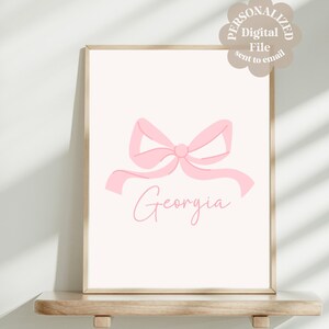 Op de afbeelding: Een printbaar digitaal bestand met een roze strik met de naam "Georgia" eronder geschreven in cursief. Het ontwerp is perfect voor een kinderkamer of babykamer.