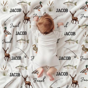 May include: A white baby blanket with a fishing and hunting theme. The blanket features illustrations of fish, ducks, deer, fishing poles, and the name "JACOB" in black. A baby in a white onesie is lying on the blanket.