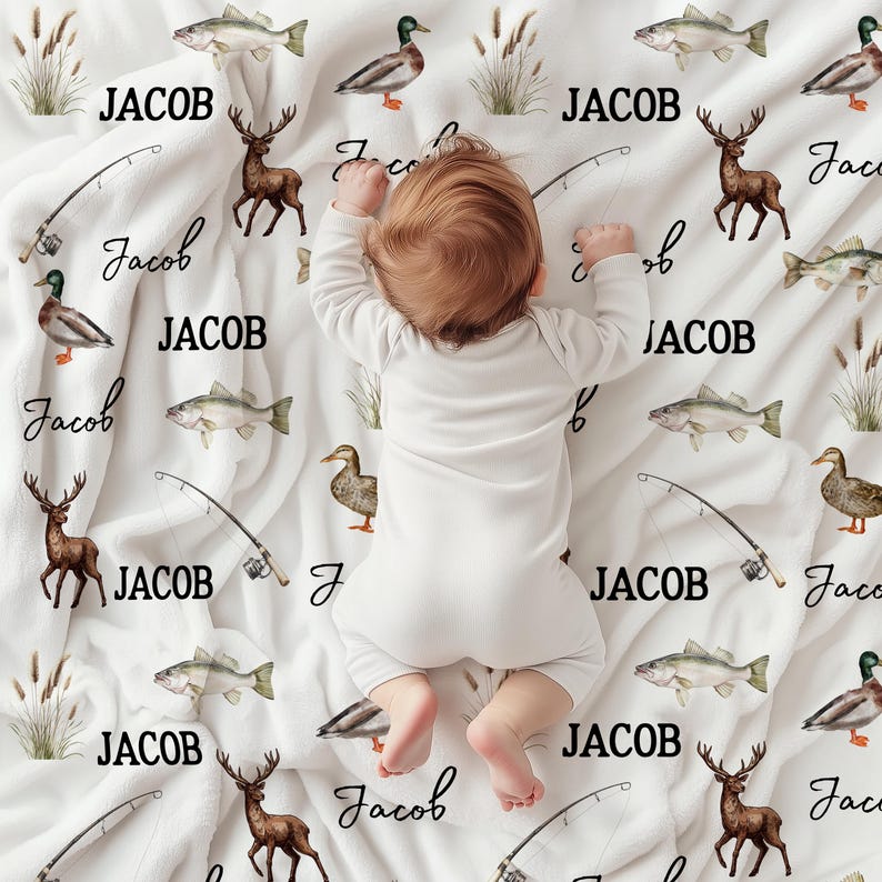 May include: A white baby blanket with a fishing and hunting theme. The blanket features illustrations of fish, ducks, deer, fishing poles, and the name "JACOB" in black. A baby in a white onesie is lying on the blanket.