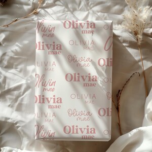 May include: White wrapping paper with a repeating pattern of the name "Olivia mae" in a soft pink color. The names are in a mix of cursive and block letters.