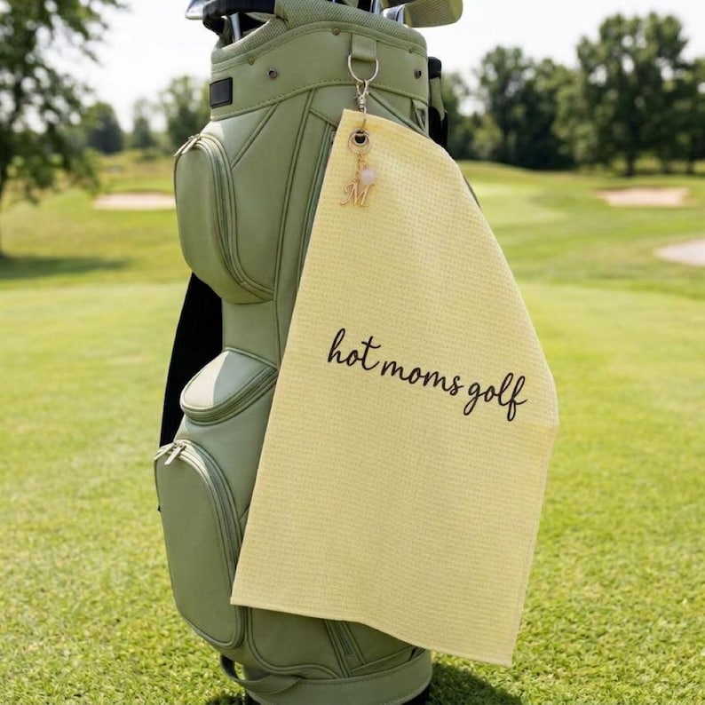 K&ouml;nnte beinhalten: Ein hellgelbes Golfhandtuch mit dem Text "hot moms golf" in schwarzer Schrift h&auml;ngt an einer Golftasche. Ein goldener Anh&auml;nger mit dem Buchstaben "M" ist am Handtuch befestigt. Die Golftasche ist hellgr&uuml;n.