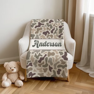 May include: A throw blanket featuring a camouflage pattern in shades of brown, tan, and cream. A horizontal white band across the center displays the name Anderson in a dark brown script font. The blanket is draped over a light-colored armchair.
