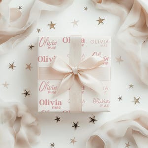 May include: A wrapped gift with a light pink ribbon and bow. The gift wrap is white with the name "Olivia mae" printed in a soft pink. Gold star-shaped confetti and sheer fabric surround the gift.