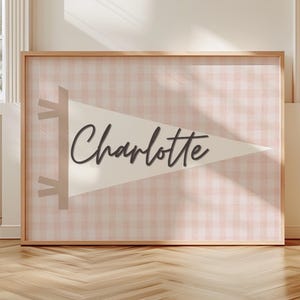Może przedstawiać: Oprawiona grafika ścienna z motywem proporczyka z napisem "Charlotte" w kursywie. Proporczyk umieszczony jest na różowo-białym wzorze w kratkę. Rama ma jasny kolor drewna.