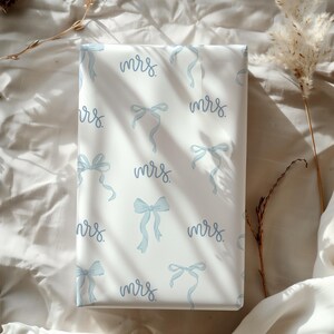 K&ouml;nnte beinhalten: Wei&szlig;e Geschenkpapier mit einem sich wiederholenden Muster aus blauen Schleifen und dem Wort "mrs." in Kursivschrift.