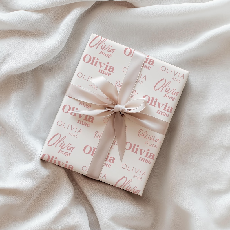 May include: A wrapped gift with a white background featuring the name "Olivia mae" in a repeating pattern in a soft pink color. The gift is tied with a matching pink ribbon and bow.