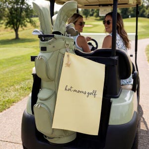K&ouml;nnte beinhalten: Eine hellgr&uuml;ne Golftasche mit passenden Schl&auml;gerhauben ist an einem Golfwagen befestigt. Ein gelbes Schild mit dem Text "hot moms golf" h&auml;ngt an der Tasche. Zwei Frauen sitzen im Wagen und tragen Sonnenbrillen.