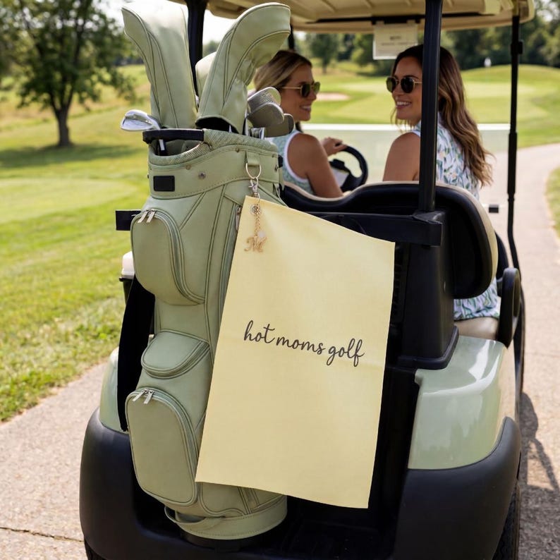 K&ouml;nnte beinhalten: Eine hellgr&uuml;ne Golftasche mit passenden Schl&auml;gerhauben ist an einem Golfwagen befestigt. Ein gelbes Schild mit dem Text "hot moms golf" h&auml;ngt an der Tasche. Zwei Frauen sitzen im Wagen und tragen Sonnenbrillen.