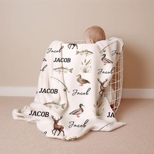 May include: A white baby blanket with a fishing theme, featuring illustrations of fish, ducks, deer, and fishing rods. The name "Jacob" is written in a cursive font across the blanket. The blanket is draped over a white wire basket.