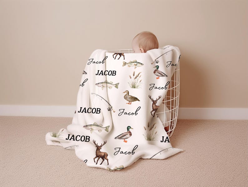 May include: A white baby blanket with a fishing theme, featuring illustrations of fish, ducks, deer, and fishing rods. The name "Jacob" is written in a cursive font across the blanket. The blanket is draped over a white wire basket.