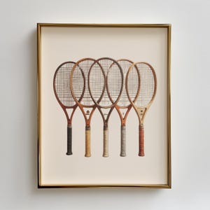 May include: A framed print featuring five vintage tennis rackets. The rackets have wooden frames and various grip colors, including black, tan, and red. The print is set against a cream-colored background and framed in gold.