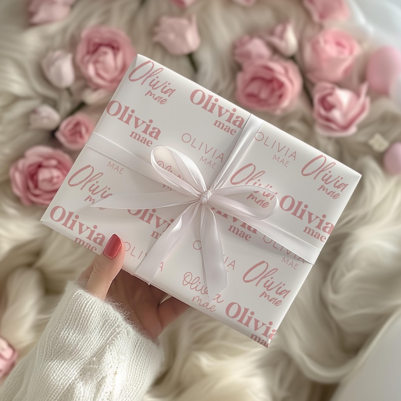May include: A wrapped gift with a white background and a pink pattern of the name "Olivia mae". The gift is tied with a white ribbon. Pink roses and a fluffy white surface are in the background.