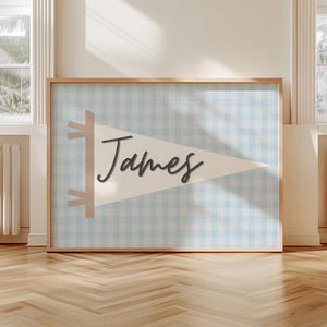 May include: A framed wall art piece featuring a light blue and white gingham pattern background. A white pennant with the name "James" in a dark script is centered. The frame is a light wood color.