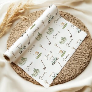 May include: A roll of white wrapping paper featuring a golf-themed design. The pattern includes golf carts, golf balls, golf clubs, and the name "James" in a handwritten style. The illustrations are in green, brown, and beige.