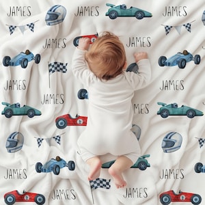 Könnte beinhalten: Eine weiße Babydecke mit Rennwagen-Motiv. Die Decke zeigt Illustrationen von blauen, roten und grünen Rennwagen, Rennhelmen, Zielflaggen und dem Namen "JAMES" in schwarzer Schrift. Geeignet für Babys.