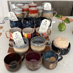 May include: A collection of handmade ceramic mugs in various colors and patterns, including blue, red, and brown. Some mugs have handwritten text and tags that read "Taylor Sips". A small bowl with a ball of yarn is also present.