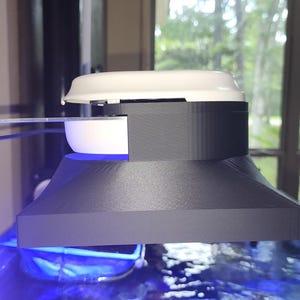 May include: A black and white aquarium light fixture with a white top and a blue light. The fixture is mounted above an aquarium, with a clear bracket attached to the side. The background shows a window with trees.