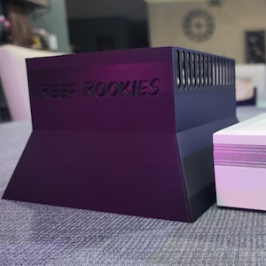 May include: Two electronic devices on a grey surface. One is a dark purple, angled box with the text "REEF ROOKIES." The other is a white box with the text "PopBloom" and silver trim.