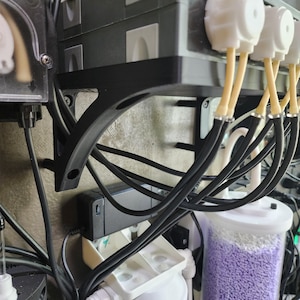 May include: Close-up of an aquarium system with various components. Black brackets support multiple black boxes with white connectors and tubing. Clear containers hold purple and white pellets. Black wires and tubes are visible.