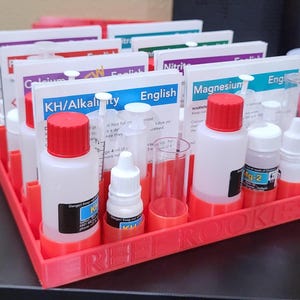May include: A red plastic tray holds several bottles, vials, and test cards for water testing. The bottles have red caps and are labeled with terms like "KH/Alkalinity" and "Magnesium." The tray is labeled "REEF ROOKIES."