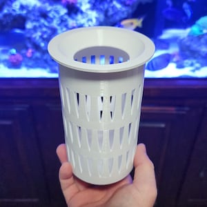 May include: A white, cylindrical aquarium filter cup with a flared top and vertical slotted sides. The cup is held in front of a blurred aquarium background with blue and green hues.