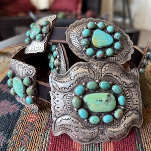 Könnte beinhalten: Ein brauner Ledergürtel mit verzierten Silberschnallen und Türkissteinen. Die Schnallen haben aufwendige Designs und sind oval und rechteckig. Die Türkissteine sind eine Mischung aus hell- und dunkelblau-grün. Der Gürtel wird auf einer farbenfrohen, gemusterten Oberfläche präsentiert.