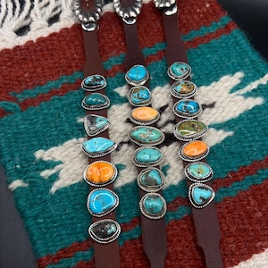 May include: Three brown leather bracelets with silver buckles and turquoise, orange, and green gemstone accents. The gemstones are set in silver bezels. The bracelets are displayed on a colorful woven textile.